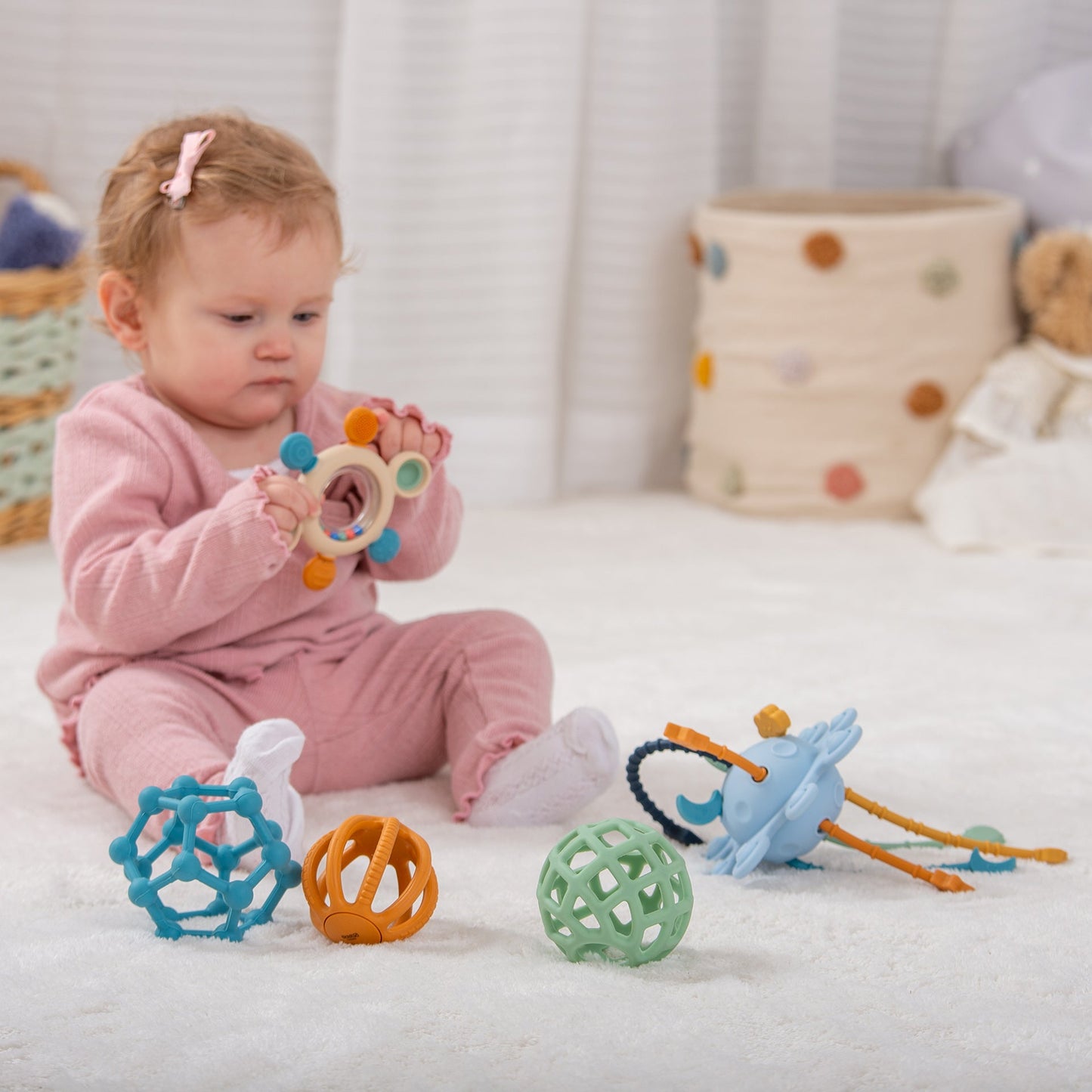 Baby playing with colorful sensory toys on soft rug, including rattle, textured balls, and teether for early development