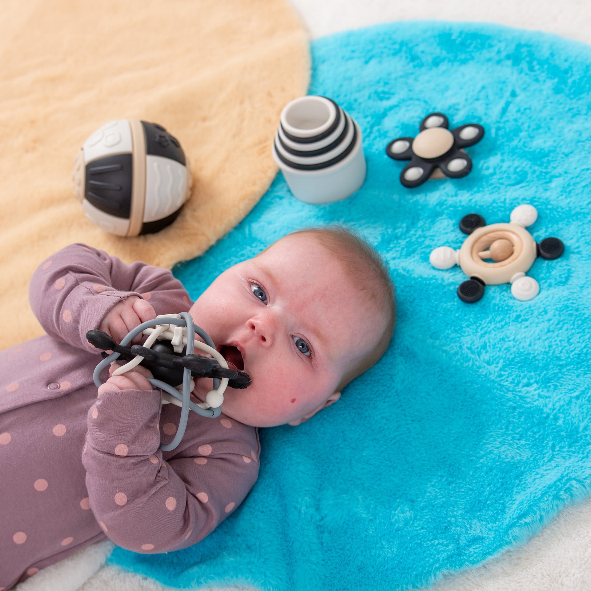 Baby chewing on teething toy surrounded by sensory baby toys on soft play mat