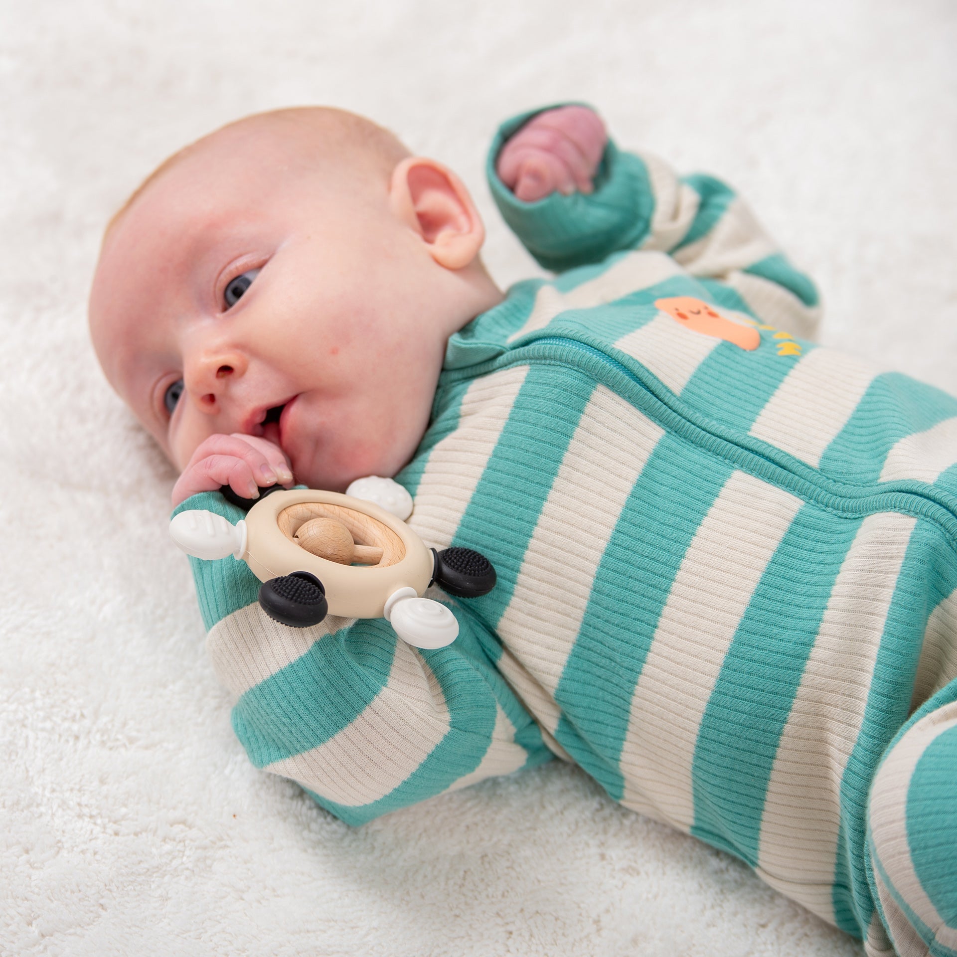 Baby chewing on wooden teething toy, wearing striped onesie on soft blanket