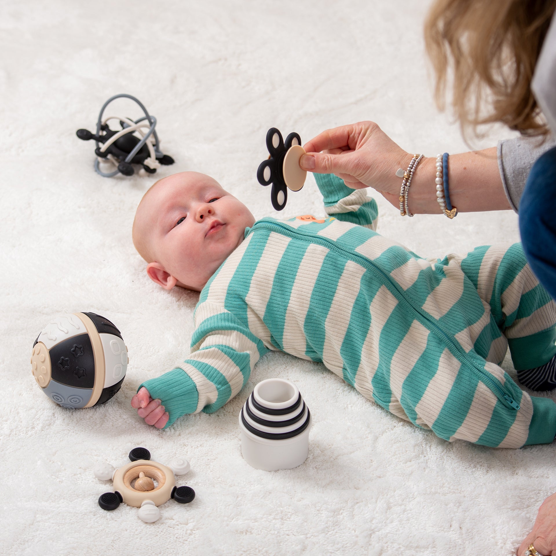 Baby in striped onesie playing with developmental toys on white rug, adult hand offering butterfly teether