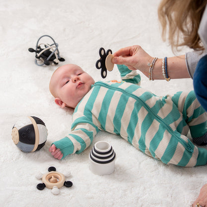 Baby in striped onesie playing with developmental toys on white rug, adult hand offering butterfly teether