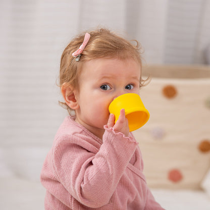 Toddler drinking from yellow sippy cup, wearing pink outfit and hair clip, ideal for baby feeding and playtime.